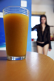 A glass of orange juice on a table with a girl in a sheer blouse in the background