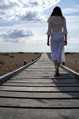 female in white lace walks on wooden walkway across shingle