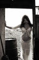 A pregnant woman in white lace stands illuminated in the doorway to an old wooden hut