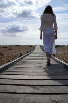 female in white lace walks on wooden walkway across shingle