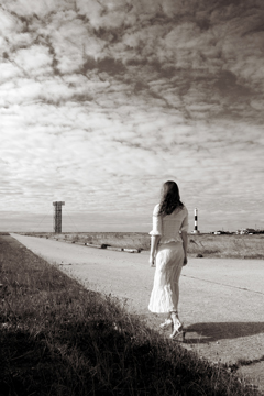 woman in white lace walks along a path with a lighthouse and shipping market in the distance