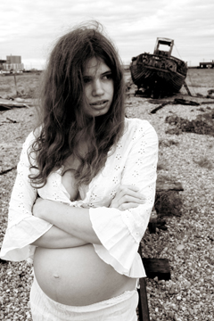 pregnant girl in white lace braces herself against a breeze with old rotting boat in background