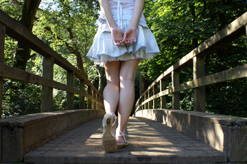 Girl in short denim skirt and sandals walks across a wooden bridge into the woods