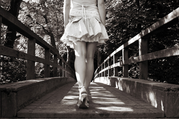Girl in short denim skirt and sandals walks across a wooden bridge into the woods